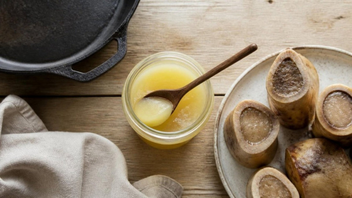 Ausgelassenes Knochenmark im Glas mit Holzlöffel, daneben Markknochen und eine Gusseisenpfanne auf einem Holztisch
