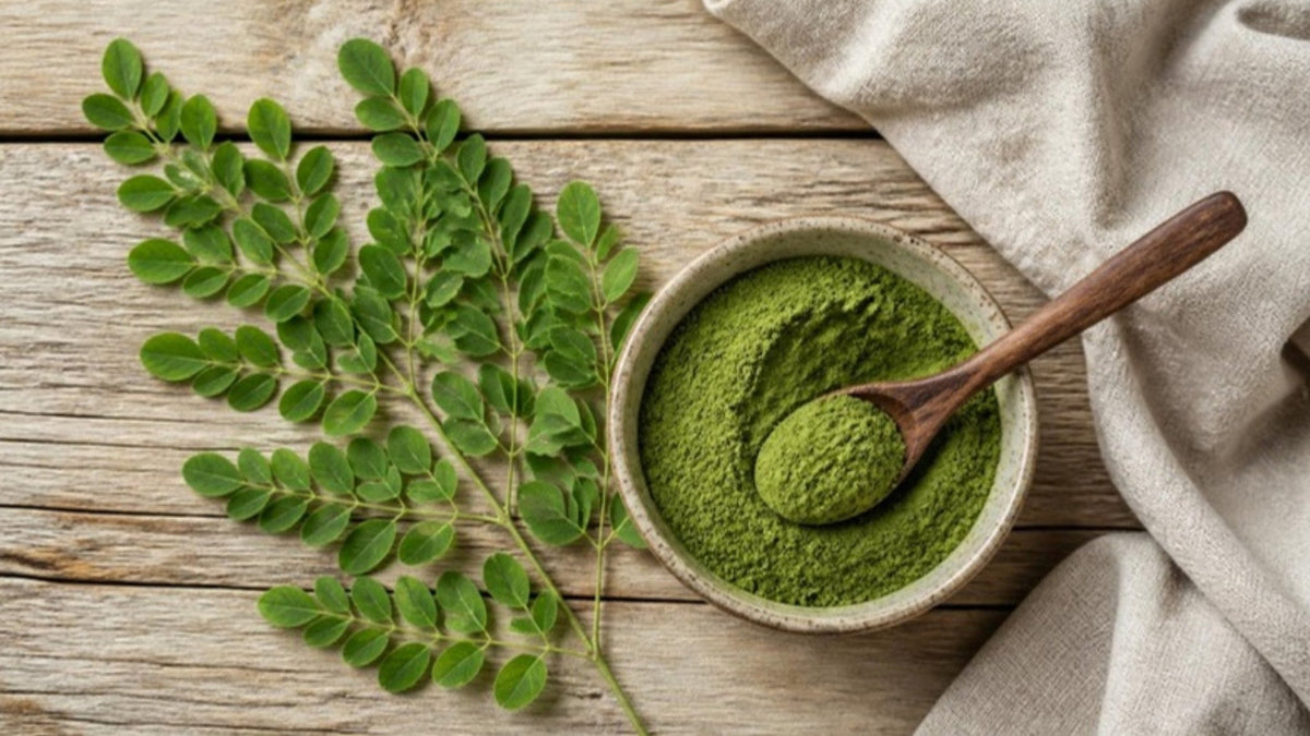 Frische Moringa-Blätter an einem Zweig neben einer Schale mit grünem Moringa-Blattpulver auf einem Holztisch