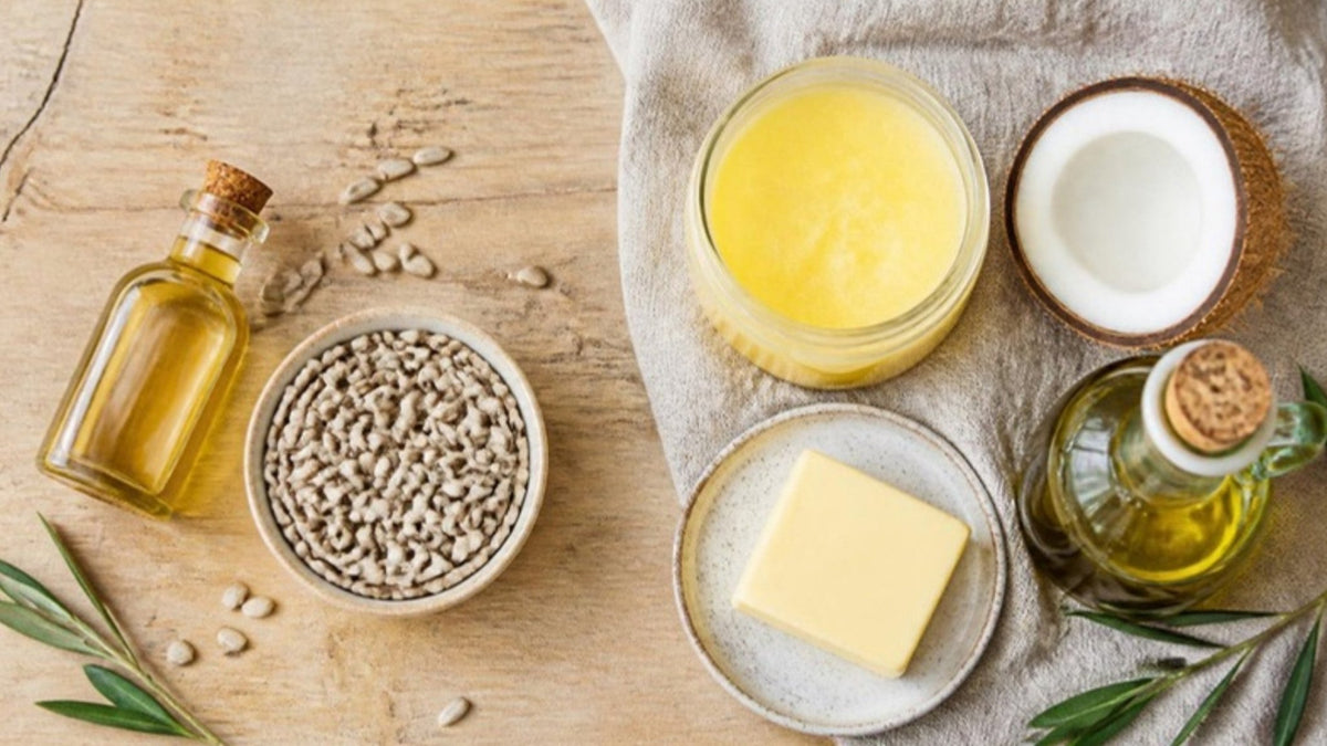 Ölflasche und Sonnenblumenkerne neben Butterschmalz, Butter, Kokosnuss und Olivenöl auf einem Holztisch