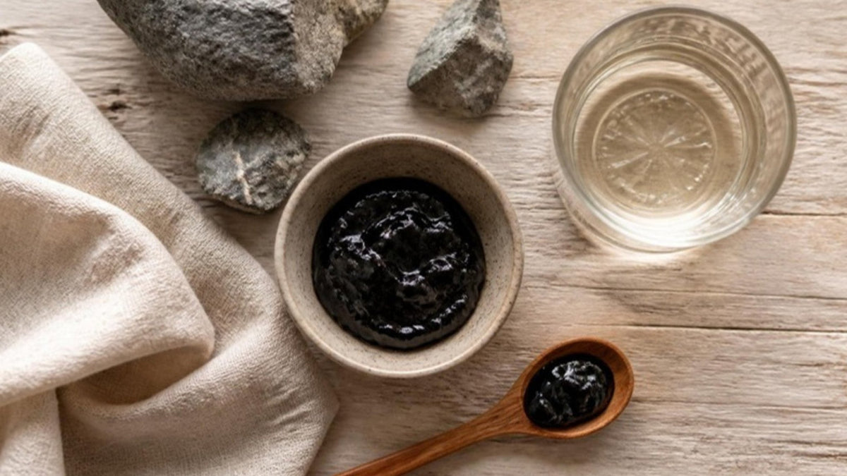 Dunkles Shilajit-Harz in einer Schale mit Löffel, Wasserglas und Steinen auf einem Holztisch